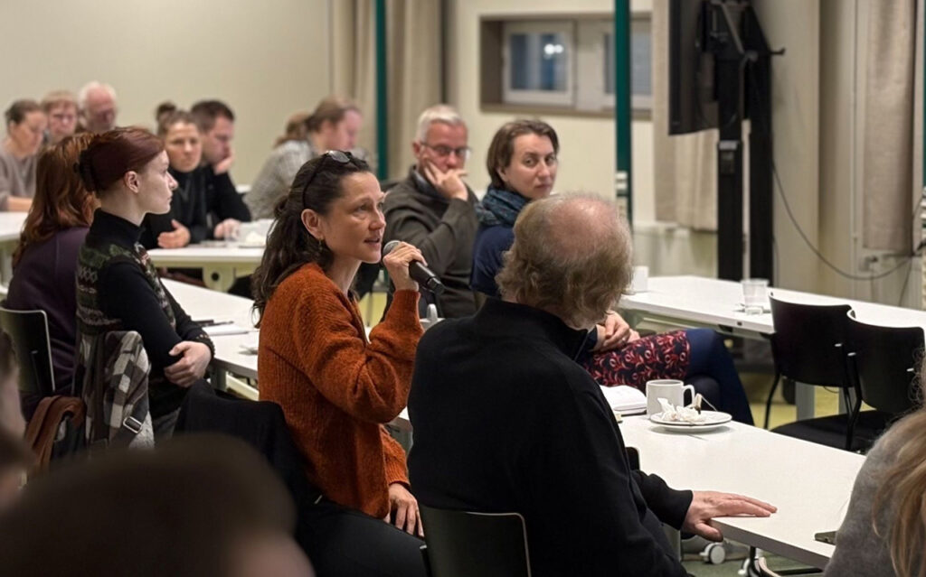 [Alt-teksti: luentosali, jossa on ihmisiä pöytien ääressä kuuntelemassa, yksi henkilö puhuu mikrofoniin.]