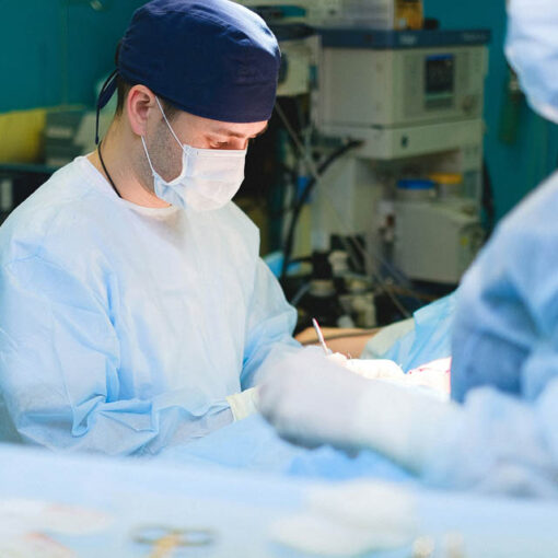 [Alt-teksi: maskiin ja suojavarusteisiin pukeutunut kirurgi työtehtävissä.]