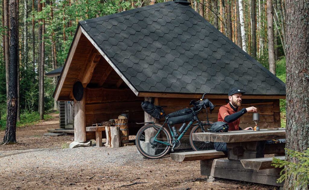 [Alt-teksti: viistokattoinen puinen laavumökki korkeiden puunrunkojen lomassa, mökin vieressä pyöräilijä istuu pitämässä taukoa pöydän ääressä.]