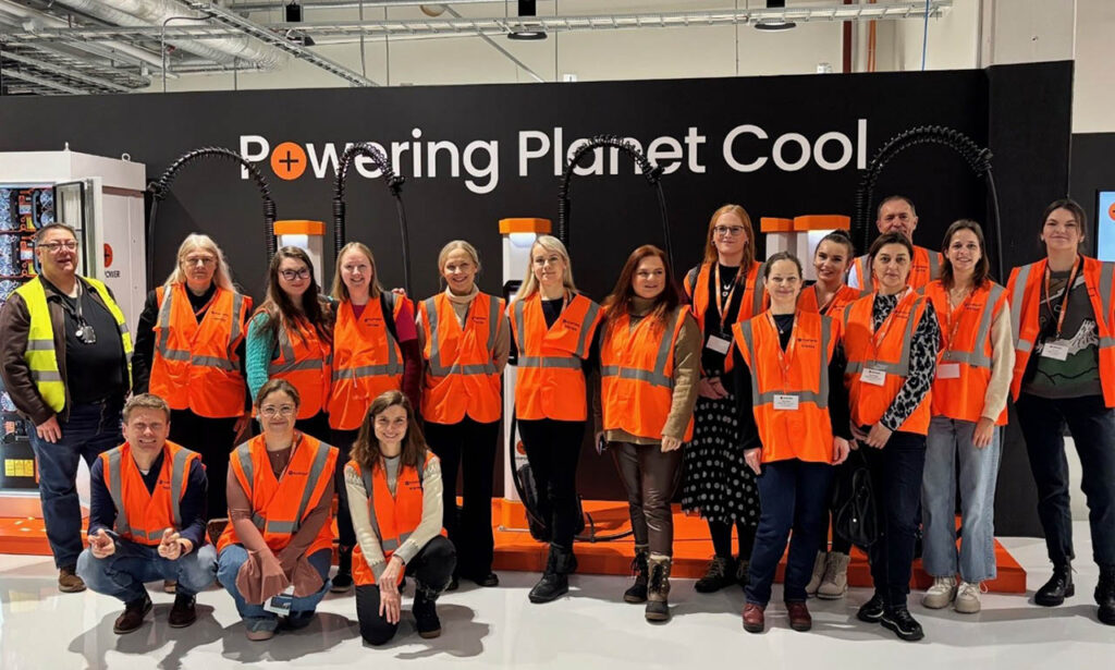 [Alt text: sixteen persons smiling in orange reflective vests in front of the wall, in the wall there is the text powering planet cool.]