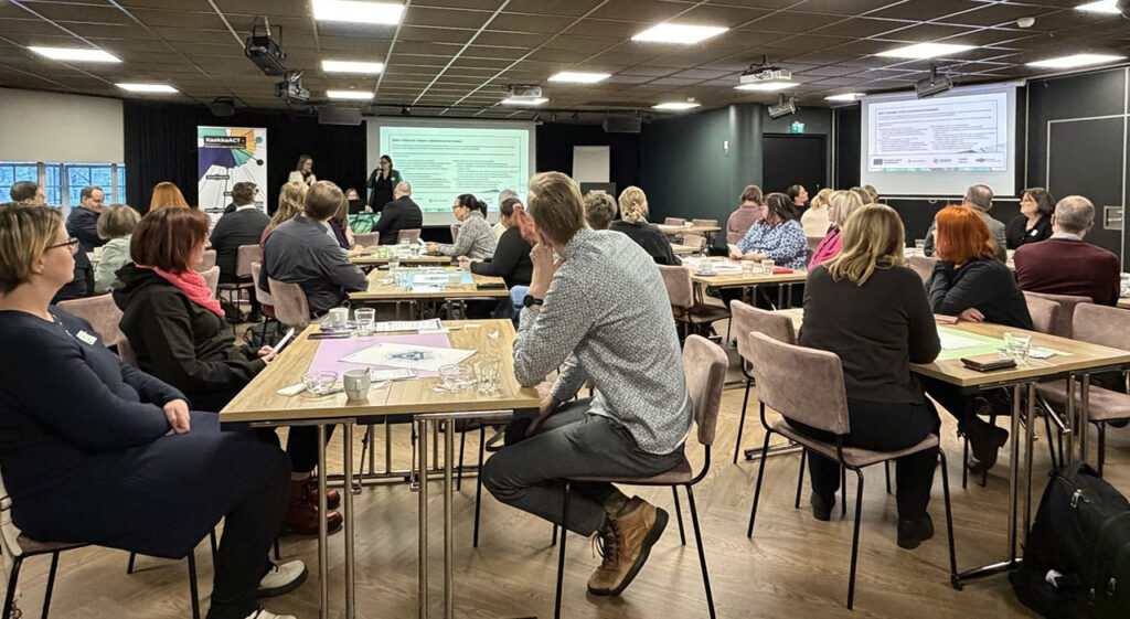 [Alt-teksti: suuri kokoustila, jossa on pöytiä ja tuoleja, pöytien ääressä ihmisiä kuuntelemassa esitystä.]