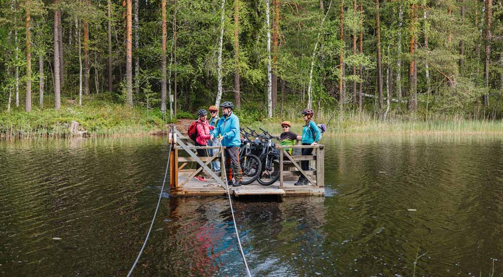 [Alt-teksti: neliskulmainen puinen lossi kulkemassa metsäjärven yli, lossin kyydissä on kypäräpäisiä retkeilijöitä sekä polkupyöriä.]