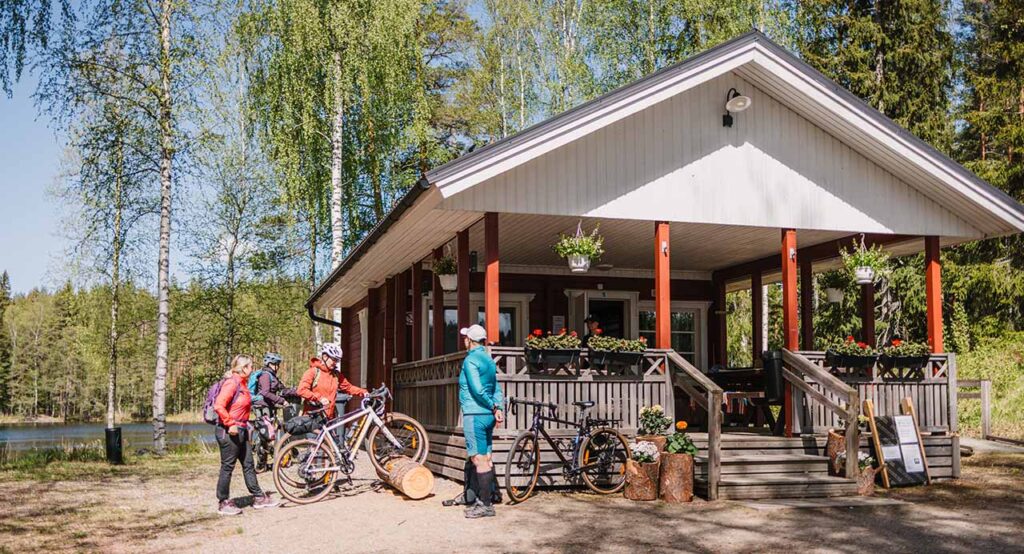 [Alt-teksti: harjakattoinen terassilla varusteltu puumökki järven rannalla koivujen katveessa, mökin vierellä pyöräilijöitä retkivarusteineen.]