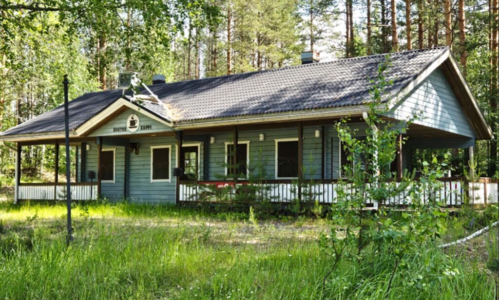 [Alt-teksti: mäntymetsän edessä ruohottuneella aukiolla seisova matala, kookas puurakennus.]