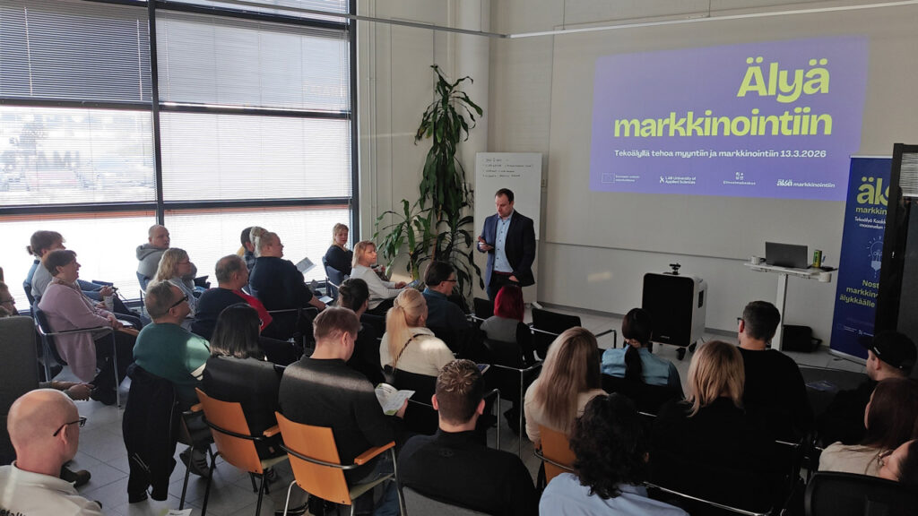 [Alt-teksti: henkilö pitämässä esitystä täydessä luentosalissa.]