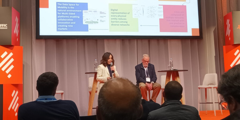 [Alt-teksti: kaksi henkilöä keskustelemassa esiintymislavalla, heidän takanaan näkyy presentaatio skriinillä.]