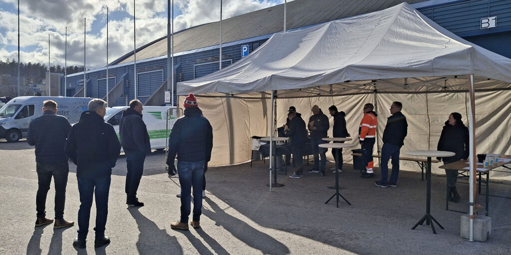 [Alt-teksti: tapahtumapaikka suuren varastorakennuksen vieressä, telttakatoksen alla seisoo väkeä pienten pöytien ääressä.]