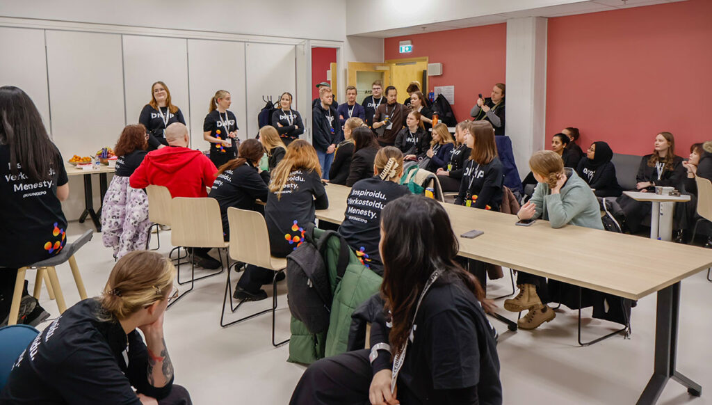 [Alt-teksti: suuri luokkahuone, jossa tungeksii opiskelijoita pöytien ääressä, seinien vierellä ja oviaukossa.]