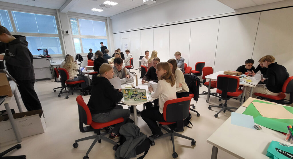 [Alt-teksti: suuri luokkahuone, jossa on parikymmentä nuorta ihmistä pöytien ympärillä työskentelemässä.]