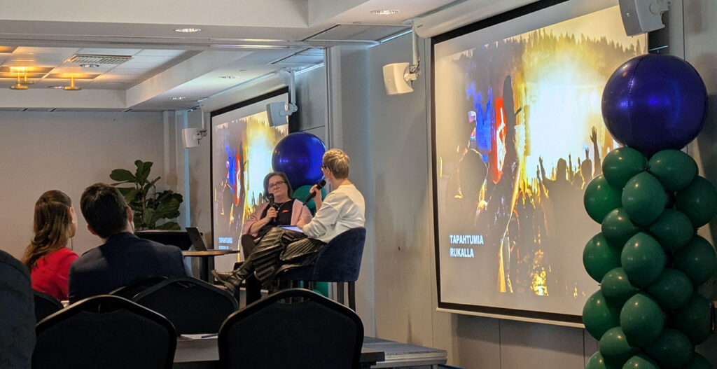 [Alt-teksti: kaksi henkilöä pitämässä esitystä yleisön edessä istuen tuoleilla ja mikrofonit käsissä.]