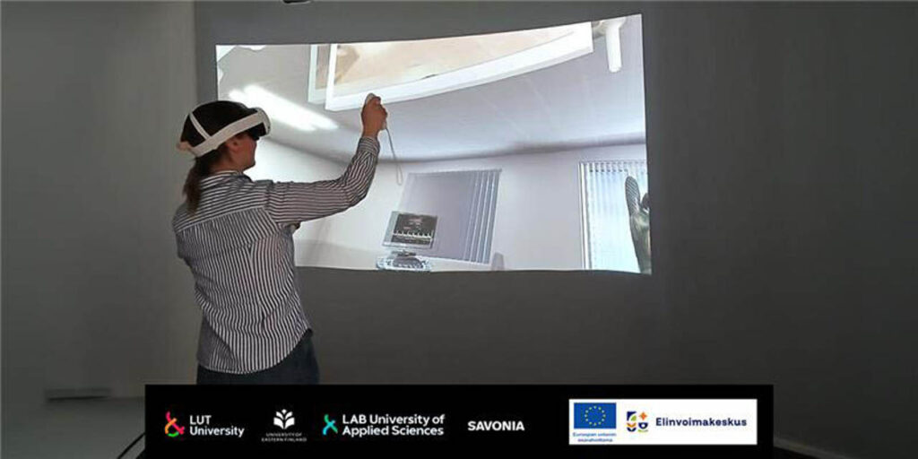 [Alt text: Henkilö seisoo neutraalisti väritetyssä huoneessa virtuaalilasit päässä ja ohjaimet kädessä. Taustalla seinälle heijastettu kuva virtuaalisesta ympäristöstä.]