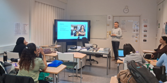Opettaja seisoo luokkahuoneessa näytön vieressä, jolla näkyy esitys. Useat oppilaat istuvat pulpettien ääressä keskittyneinä ja tekevät muistiinpanoja.