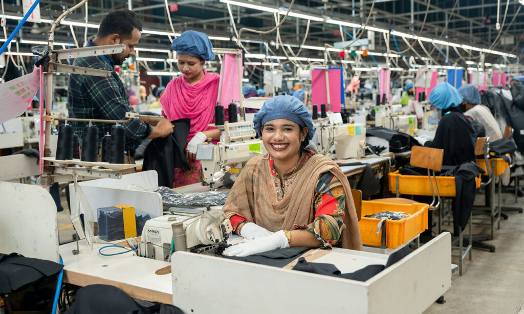 [Alt text: A smiling woman in a factory with machines and fabrics.]