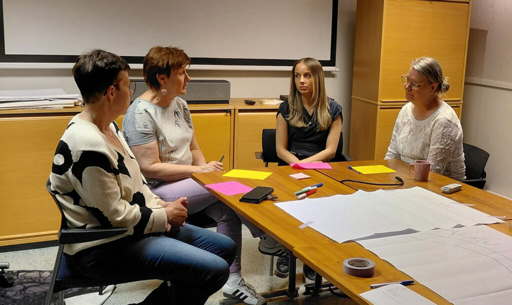[ALT-teksti: Neljä henkilöä istuu pöydän ääressä. Pöydällä on iso valkoinen paperi ja muistiinpanovälineitä.]