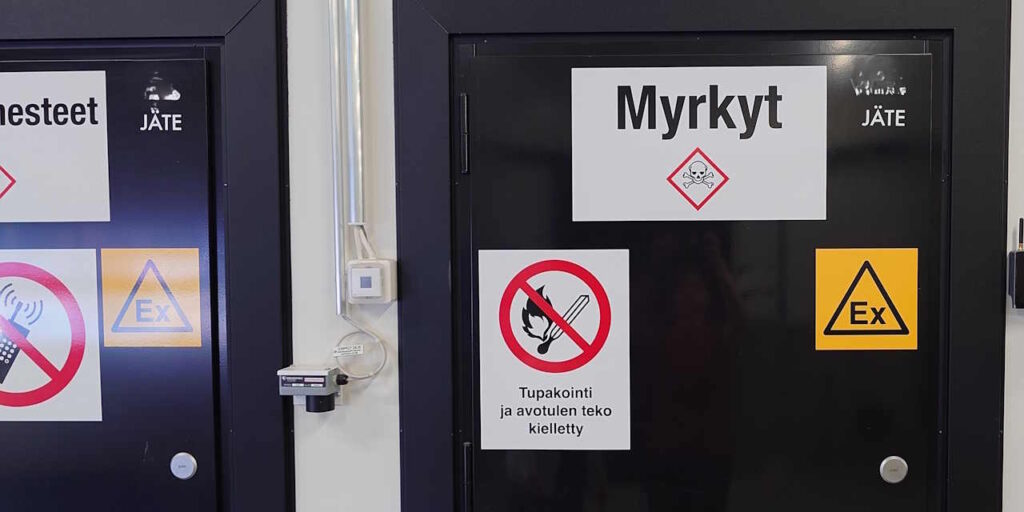Musta ovi, jossa varoitus- ja kieltokylttejä. Kylteissä lukee "Myrkyt" ja "Tupakointi ja avotulen teko kielletty".