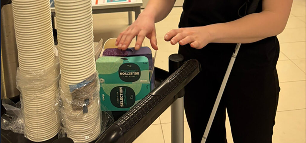 [Alt-teksti: henkilön kädet tunnustelevat teepussipakkausta, pakkauksen vieressä on kertakäyttömukeja sekä kuumajuoma-automaatti.]