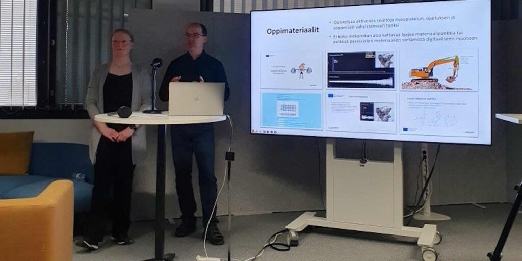 Suuri näyttö, jossa esillä esimerkkejä oppimateriaaleista. Näytön vierellä kaksi puhujaa.