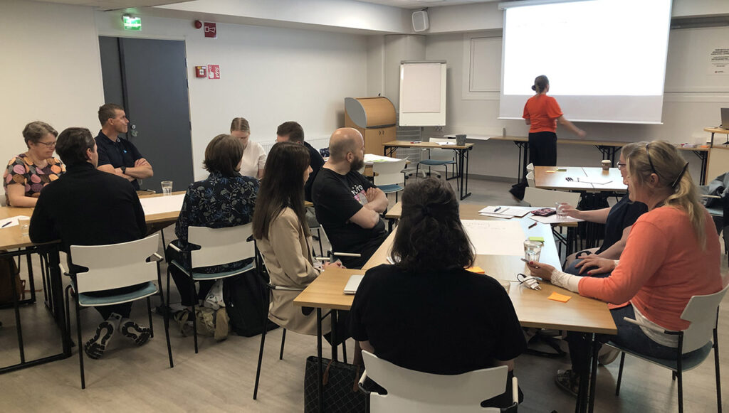 [Alt-teksti: toistakymmentä henkilöä istumassa pitkien pöytien ääressä seminaaritilassa, huoneen edessä henkilö valmistautuu pitämään esitystä.]