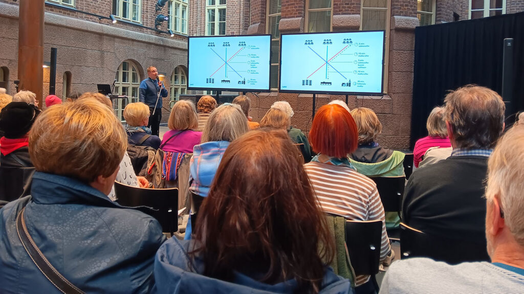 [Alt-teksti: suuren kivirakennuksen sisäpiha, jonne on kokoontunut ihmisiä, kivimuurin edessä on kaksi suurta näyttötaulua sekä henkilö pitämässä esitystä.]