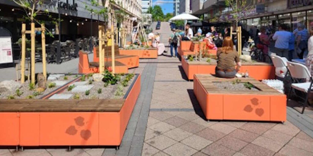 Kaupunkiaukio, jossa oransseja laatikoita, joissa kasvaa pieniä puita ja kasveja.
