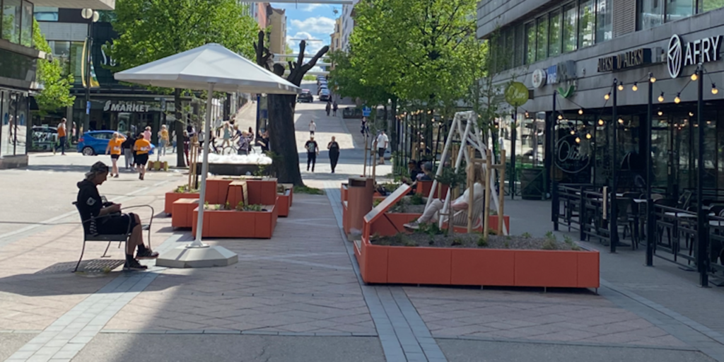 Kaupunkiaukio, jossa oransseja kasvilaatikoita, aurinkovarjo ja ihminen istumassa varjon alla.