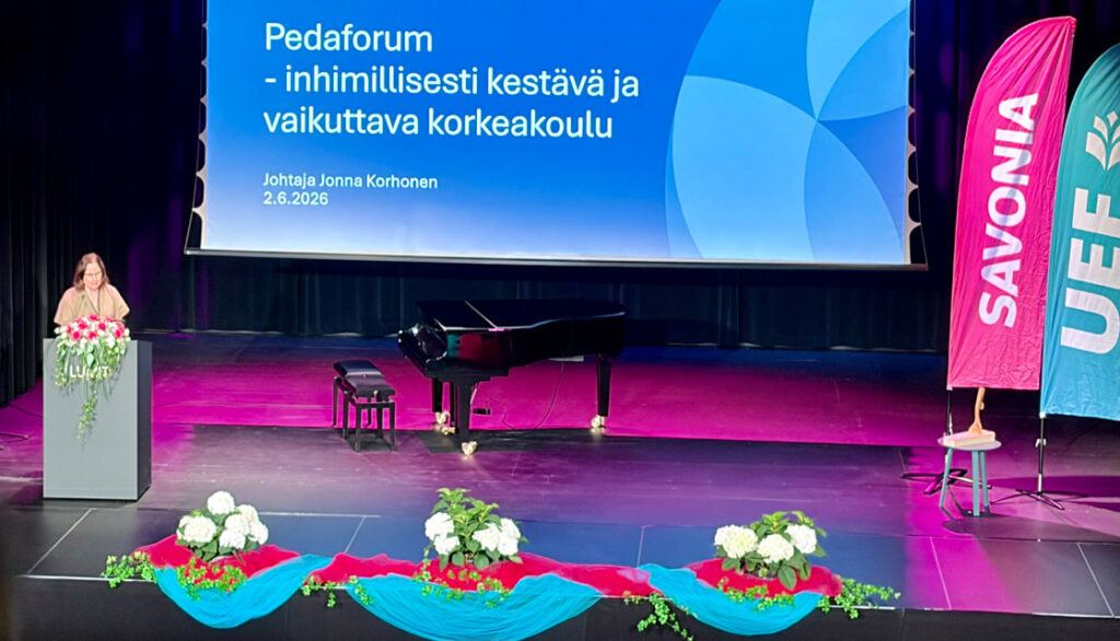 Esiintymislavan näytöllä teksti "pedaforum - inhimillisesti kestävä ja vaikuttava korkeakoulu. Puhujapöntössä ihminen, etualalla isot kukka-asetelmat.