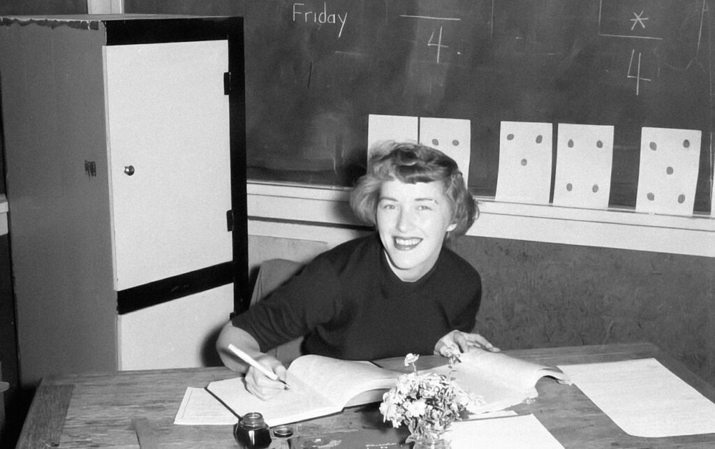 [Alt-teksti: mustavalkoinen kuva noin 1950-luvulta, leveästi hymyilevä nuori nainen kirjoittamassa merkintöjä kirjaan papereiden peittämällä työpöydällä.]