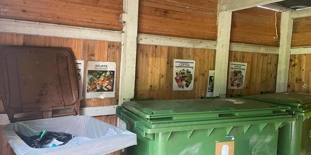 A covered residential waste collection point with three large waste containers, Waste sorting posters are mounted on the wooden wall behind the containers.